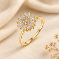 Bague au fini doré avec motif rayonnant de pierres claires, présentée sur fond ivoire et tissu crème